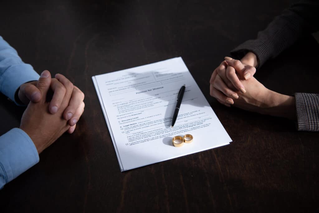 photo of divorce papers with a pen and two rings on top of them. On one side of the paperwork is a woman's folded hands, on the other a man's folded hands.